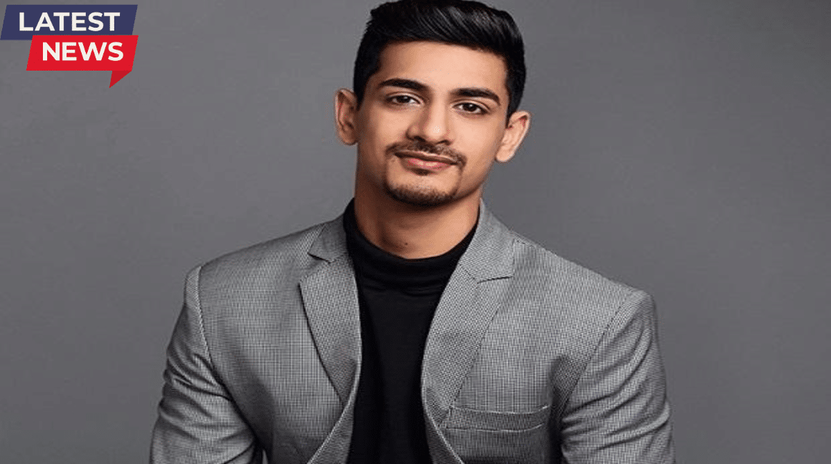 Portrait of a young man wearing a grey blazer over a black turtleneck, looking confidently at the camera with a clean studio background and “Latest News” label.