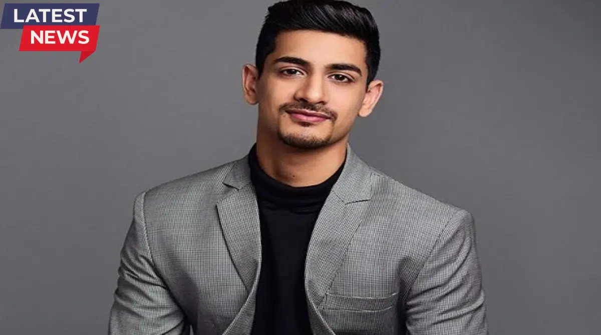 Portrait of a young man wearing a grey blazer over a black turtleneck, looking confidently at the camera with a clean studio background and “Latest News” label.