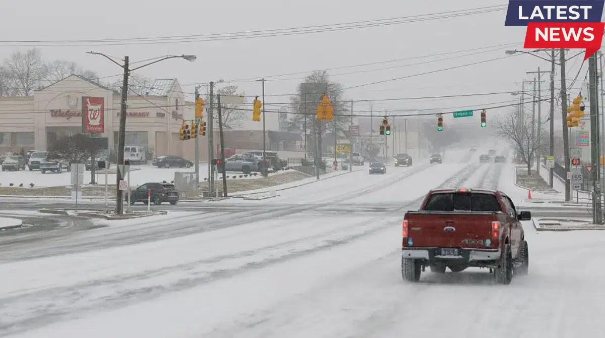 Snow-covered road with vehicles driving slowly during a winter snowstorm in a U.S. town as traffic lights glow through heavy snowfall.